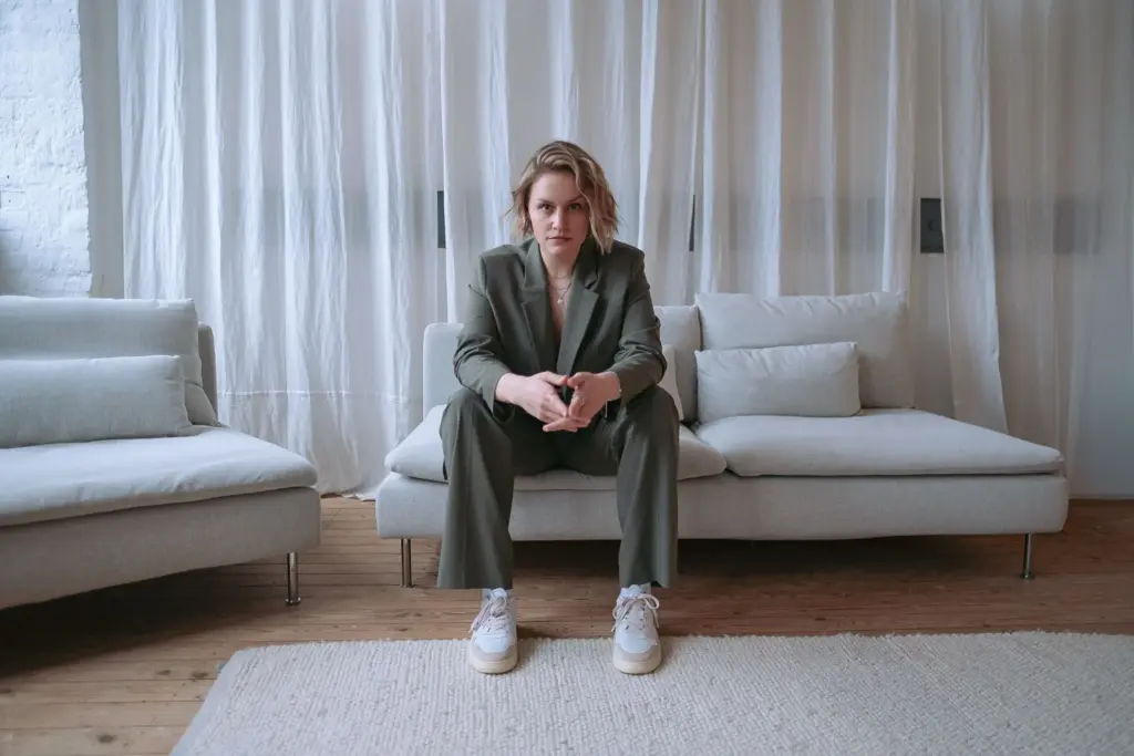 Maischa Pingel sitzt mit einem olivfarbenen Blazer auf einer beigen Couch vor einem beigen Vorhang.