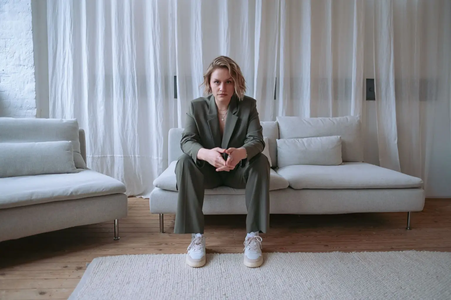 Maischa Pingel sitzt mit einem olivfarbenen Blazer auf einer beigen Couch vor einem beigen Vorhang.